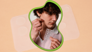 Teenager boy holding sheet of pimple patches in left hand whilst applying one patch to his face with his right hand. He has dark curly hair and is wearing a pink collared shirt.
