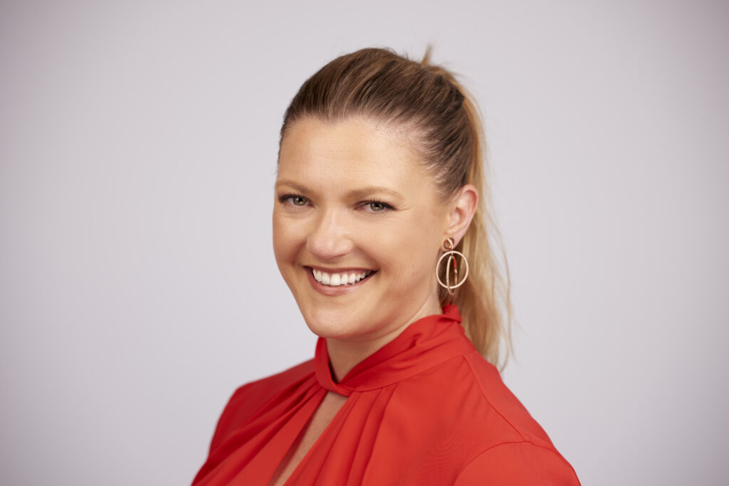 Lisa Solomons is smiling at the camera, wearing a orange shirt and has long brown hair up in a pony tail